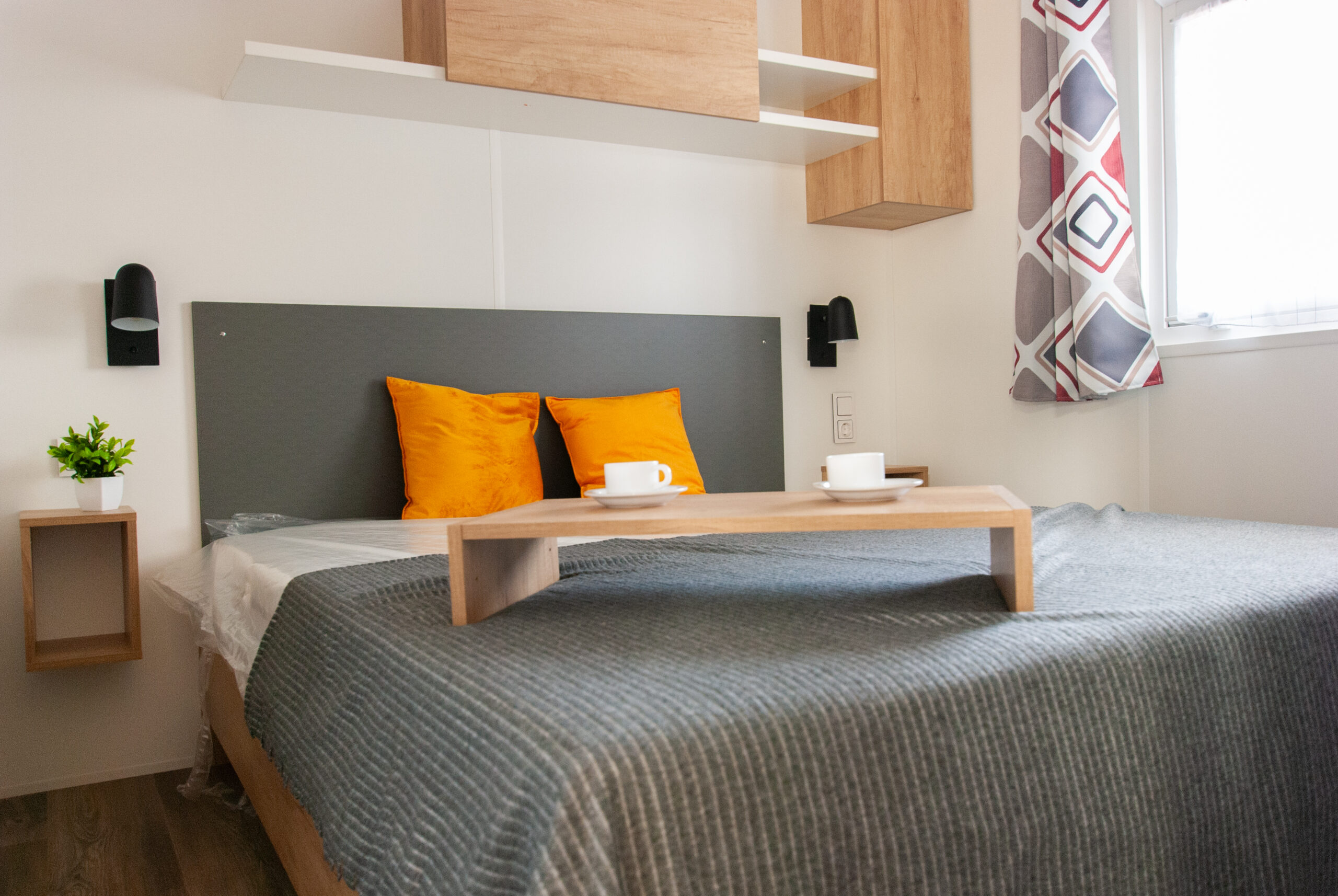 Dormitorio principal en una casa móvil Rapid'home Elite 902 en venta en las Landas, con una cama doble con mantas grises y almohadas naranjas.