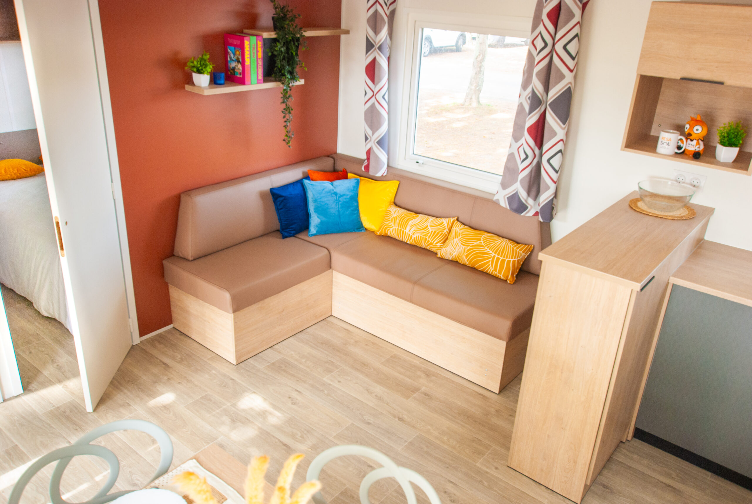 Salon lumineux du mobil home Rapid'home LO872 à vendre dans les Landes, avec grand canapé et oreillers pour 4 personnes, bénéficiant de lumière naturelle grâce à la fenêtre et à la baie vitrée d'entrée